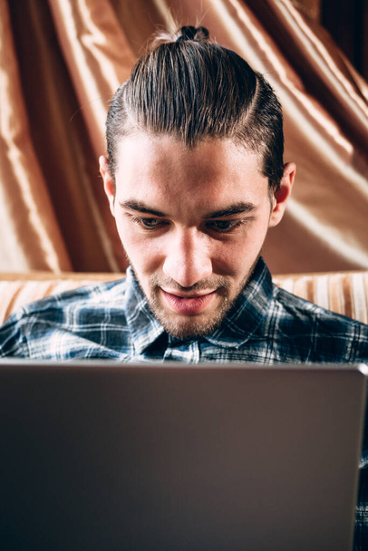 Un apuesto joven freelancer con una camisa azul trabaja en una laptop plateada en casa. Trabajo remoto durante la cuarentena. - Foto, Imagen