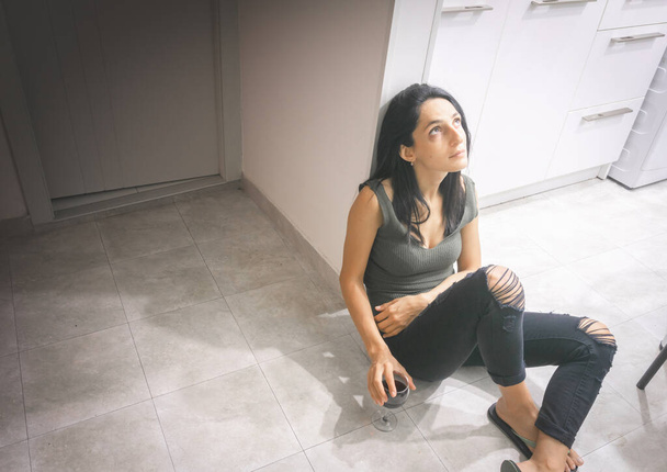 Jovem bela mulher caucasiana senta-se no chão da cozinha e olha para a luz com rosto triste e cansado - Foto, Imagem