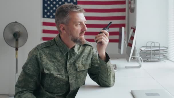 Langzaam zijaanzicht schot van een bebaarde mannelijke legerofficier in militair uniform zittend aan het bureau en pratend op walkietalkie in functie - Video