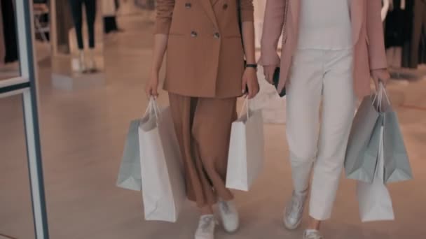 Slowmo tracking con tilt up di giovani donne felici che trasportano borse della spesa che escono dal negozio di vestiti nel centro commerciale e chatta - Filmati, video