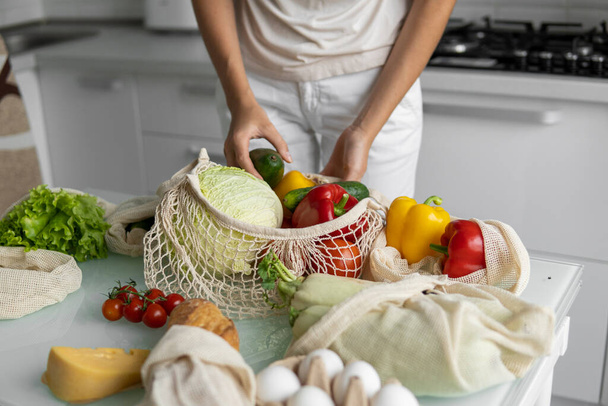 Woman came back from a market and unpacks a reusable grocery bag full of vegetables on a kitchen at home. Zero waste and plastic free concept. Girl is holding mesh cotton shopper with vegetables. - Фото, изображение