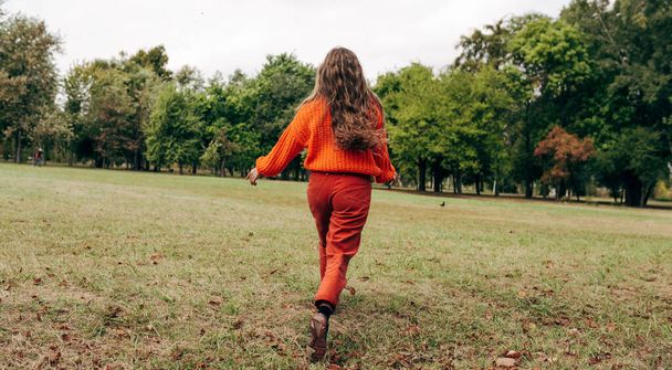 Rear view of an young woman walking with blowing long blonde hair wearing an orange sweater, posing on the nature background. Pretty female playing with her hair outdoor in the park.  - Foto, Imagem