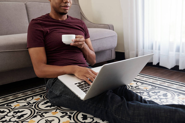 Gülümseyen genç siyah adam bir fincan kahve içiyor ve sabahları e-postaları kontrol ediyor. - Fotoğraf, Görsel