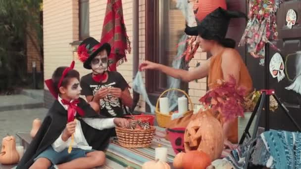 魔女の帽子をかぶった若い母親と、ハロウィーンのために装飾された家の玄関に座っている怖いメイクや衣装を着た2人の男の子の手持ち撮影。子供たちはトリックや治療後にキャンディーを食べています - 映像、動画