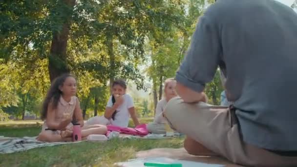 Sledování úrovně záběru mužského učitele sedí se zkříženýma nohama na přikrývce v parku a mluví se skupinou školáků, zatímco jedí jablka a pozorně ho poslouchají - Záběry, video