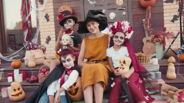 Handheld-Portraitaufnahme einer jungen Frau mit Hexenhut und drei Kindern in Halloween-Kostümen, die auf den Stufen vor der Veranda des Hauses sitzen und für die Kamera lächeln - Filmmaterial, Video
