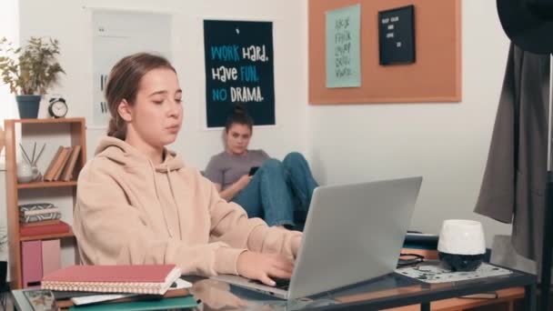 Sledování záběru veselá dívka sedí za stolem na kolejích a psaní na notebook, pak se ptát od svého spolubydlícího relaxaci na posteli v pozadí - Záběry, video
