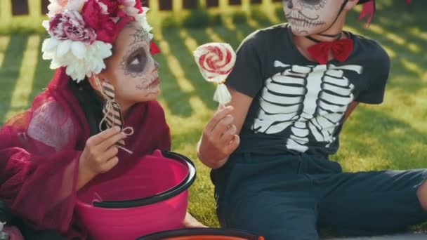 Наклоните снимок детей в костюмах на Хэллоуин, сидящих на обочине в солнечный день и смотрящих на конфеты, которые они собирали за сладостями. Мальчик дает большой леденец девушке - Кадры, видео