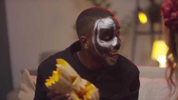 Slowmo mittlere Aufnahme eines glücklichen schwarzen Mannes in Halloween-Schminke, der einen geschnitzten Kürbis hält und ihn einer fröhlichen jungen Frau auf einer Hausparty zeigt - Filmmaterial, Video