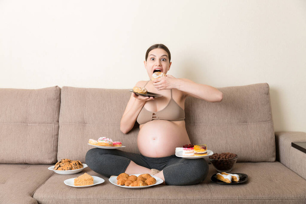 Hongerige zwangere vrouw die op de bank zit, eet veel ongezond voedsel zoals donuts, gebak, chocolade cornflakes, croissants en koekjes. Zoete hunkering tijdens de zwangerschap concept. - Foto, afbeelding