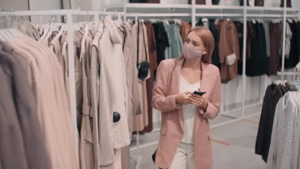 Description: Slowmo medium shot of young woman in face mask typing on mobile phone and shopping in clothes store. She is taking sweater off garment rack and walking away - Footage, Video