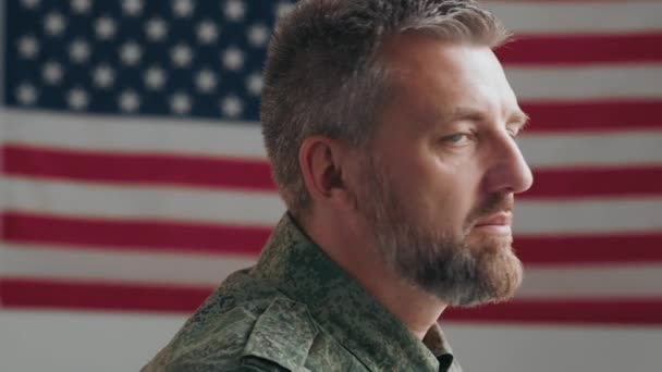 Portret van mannelijke legerveteraan van middelbare leeftijd in militair uniform die zich omdraait naar camera en salueert terwijl hij voor de vlag van de VS poseert - Video