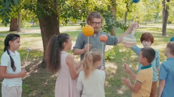 Sledování záběru starostlivého učitele vědy s vousy a skupiny zvědavých dětí držících modely planet na tyčích a učení se o sluneční soustavě v parku v letní den - Záběry, video