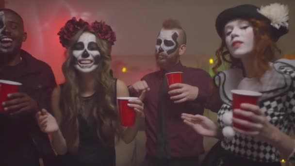 Kézi portré boldog fiatal férfiak és nők halloween jelmezek kezében műanyag poharak tele alkohollal és tánc ház party - Felvétel, videó
