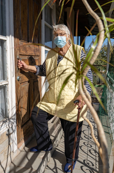 年上の女性は保護顔マスクを着用します。老婦人は屋外のドアの横にあります。家にいて、コヴィト-19の間にマスクをつけて - 写真・画像