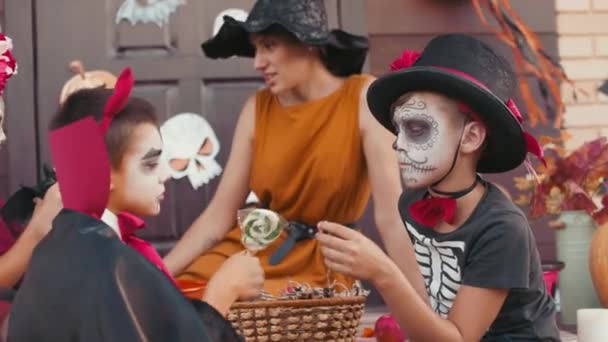 Handgehaltene Aufnahme einer jungen Frau mit Hexenhut, die auf der Veranda des Hauses sitzt und mit drei Kindern in Halloween-Kostümen plaudert, während sie Süßigkeiten essen, nachdem sie getrickst oder behandelt haben - Filmmaterial, Video