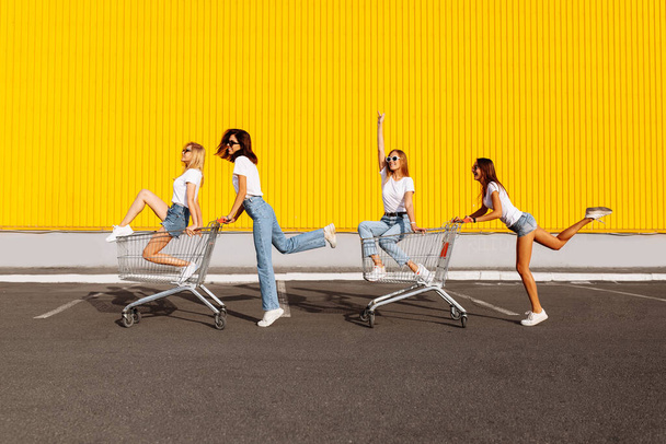 Group of happy young people having fun on shopping carts. Young beautiful women having fun and riding trolleys on a sunny summer day against the background of the yellow wall of the shopping mall - Φωτογραφία, εικόνα