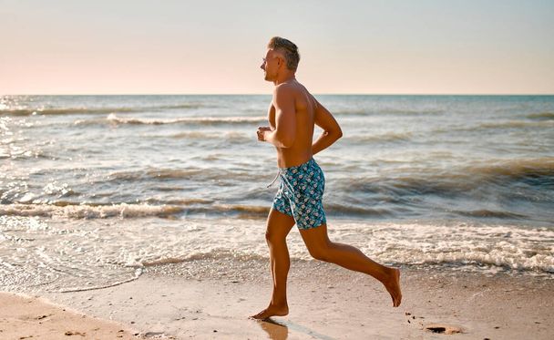 Handsome athletic man in swimming trunks running on the beach. Morning run. Healthy lifestyle concept. - Photo, Image