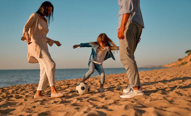 Happy family concept. Young attractive mother, handsome father and their little cute daughter having fun together and playing football on the beach. - Foto, imagen