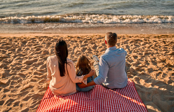 Glückliches Familienkonzept. Junge attraktive Mutter, schöner Vater und ihre kleine süße Tochter sitzen zusammen am Strand. - Foto, Bild