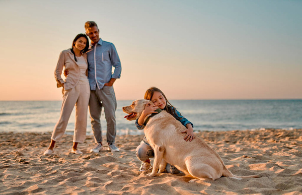 Concepto familiar feliz. Joven madre atractiva, padre guapo y su pequeña hija linda de pie juntos en la playa con el perro. - Foto, imagen