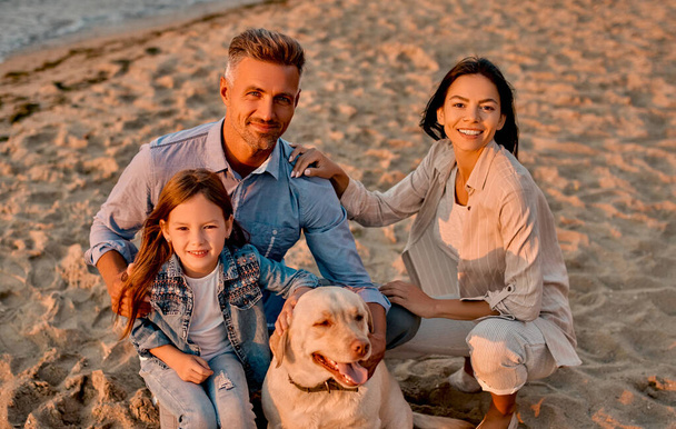 Happy family concept. Young attractive mother, handsome father and their little cute daughter sitting together on the beach with dog. - Fotografie, Obrázek