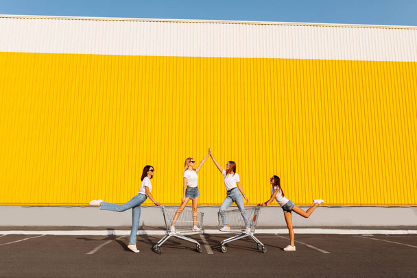 Group of happy young people having fun on shopping carts. Young beautiful girlfriends rush on a cart on a sunny summer day against the background of a yellow wall of a shopping center - Fotoğraf, Görsel