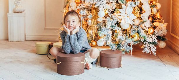 Little girl with gift box near Christmas tree on Christmas eve at home. Young kid in light bedroom with winter decoration. Christmas New Year december time for celebration concept Banner - Photo, Image