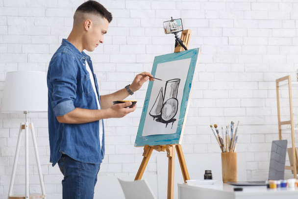 Thuisonderwijs, hobby tijdens quarantaine. Millennial man tekent vormen in beeld met verf en horloges les op smartphone in de woonkamer - Foto, afbeelding