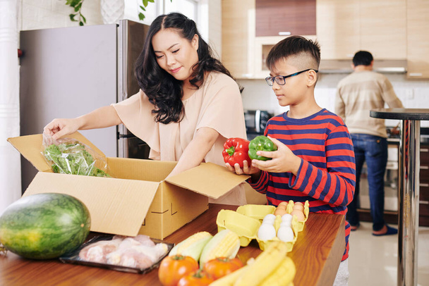 Asiatische Mutter und Sohn packen Kiste mit frischen Lebensmitteln am Küchentisch aus - Foto, Bild