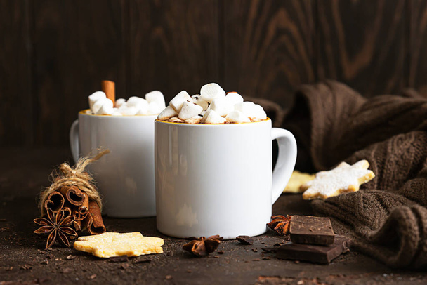 Two white ceramic cups with hot cocoa or chocolate with marshmallow and anise, cinnamon and bitter chocolate on dark stone background. - Fotoğraf, Görsel
