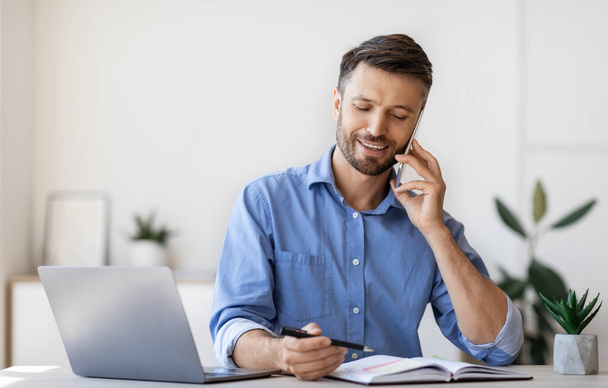 Улыбающийся бизнесмен делает заметки, разговаривая по телефону в офисе - Фото, изображение