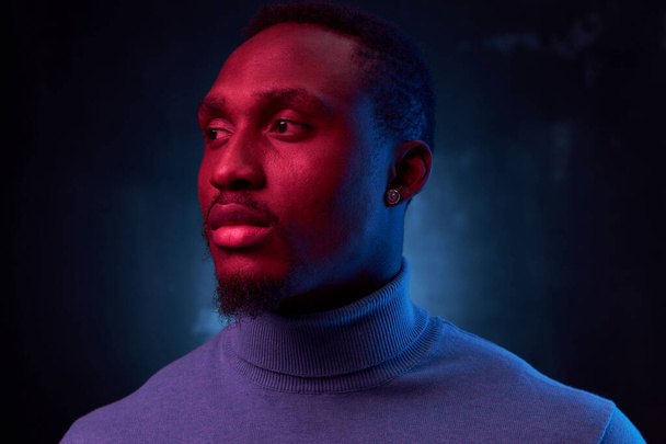 portrait photo on black background with neon light on dark-skinned handsome man in a blue turtleneck who looks away - Фото, изображение