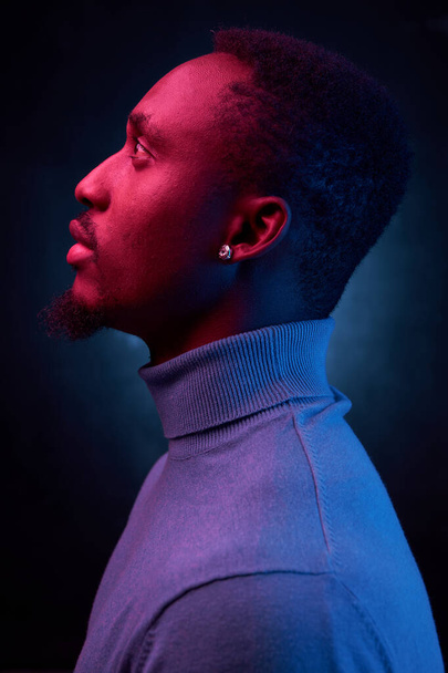 portrait photo on a dark background with neon light on a dark-skinned handsome man in a blue turtleneck standing in profile - Foto, Imagem