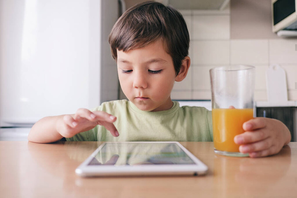 Egy helyes fiú ül az asztalnál, tablettát használ és gyümölcslevet iszik.. - Fotó, kép