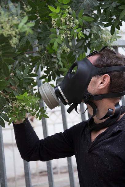 Όμορφος άντρας με προστατευτική μάσκα αερίου, έννοια προστασίας από ιούς. Κρατώντας ένα λουλούδι στο χέρι του και μυρίζοντας μέσα από μια μάσκα αερίου - Φωτογραφία, εικόνα