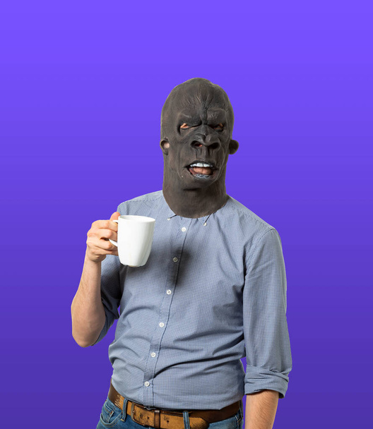 Man wearing gorilla mask holding coffee mug on isolated blue background. Medium shot.  - Photo, Image