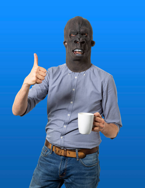ゴリラマスクの男が親指を立ててコーヒーカップを持っている。隔離された青の背景。中射.  - 写真・画像