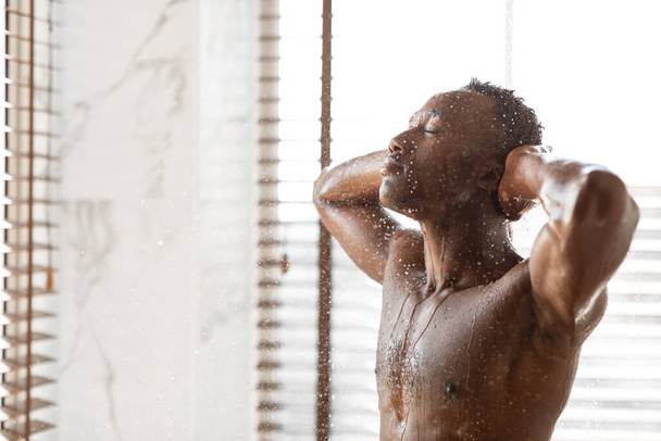 Розслаблений афроамериканський чоловік приймає душ стоячи в сучасній ванній
 - Фото, зображення