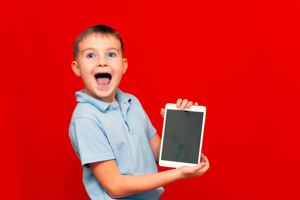 Smějící se dítě s tabletou. Chlapec ukazuje prázdné tablety na červeném pozadí - Fotografie, Obrázek