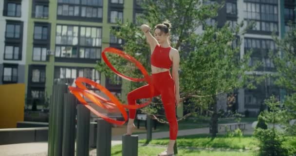 Kırmızılı genç kadın jimnastikçi apartman bahçesinde kurdele ile spor yapıyor, jimnastikçi rezidans alanında akrobatik egzersizler yapıyor, şehir ortamında fitness, 4k 120p Prores karargahı - Video, Çekim
