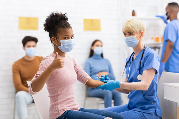 Schwarzes Mädchen gestikuliert Daumen hoch und erhält Coronavirus-Impfstoff im Krankenhaus - Foto, Bild