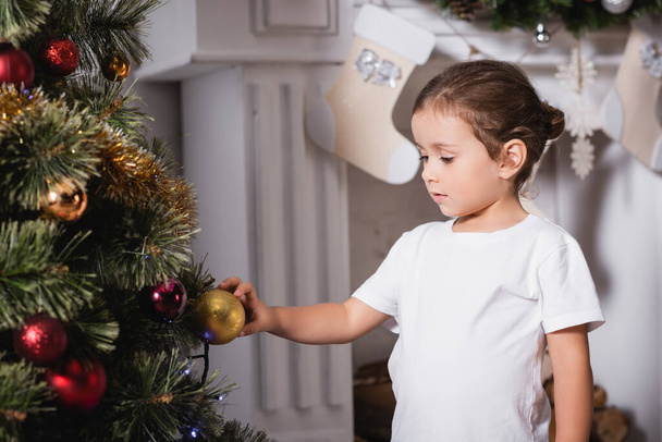 Girl standing near fireplace and touching christmas ball on festive pine at home - Φωτογραφία, εικόνα
