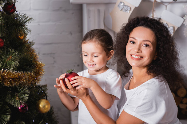 Mother and daughter with christmas ball near festive pine at home - Fotoğraf, Görsel
