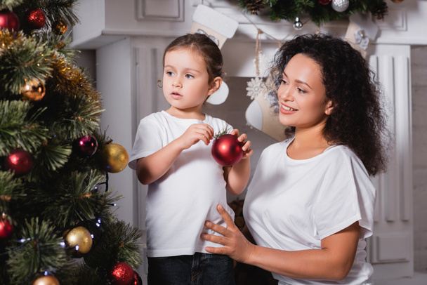 Madre e hija con bola de Navidad de pie cerca de pino festivo y chimenea - Foto, Imagen