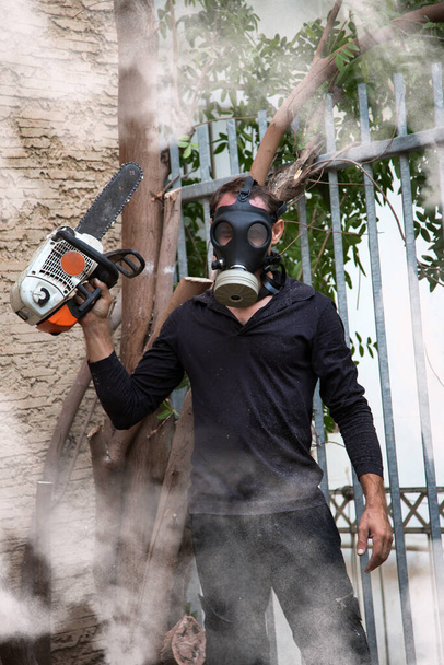 Άντρας με μάσκα αερίου στη μέση του καπνού του αλυσοπρίονου. Επικίνδυνη δουλειά. Δυνατό αλυσοπρίονο. Ο ξυλοκόπος κρατάει αλυσοπρίονο. Εξοπλισμός ξυλοκόπων για κηπουρούς. Εργασία στον κήπο κοντά στο σπίτι κατά τη διάρκεια καραντίνας.  - Φωτογραφία, εικόνα