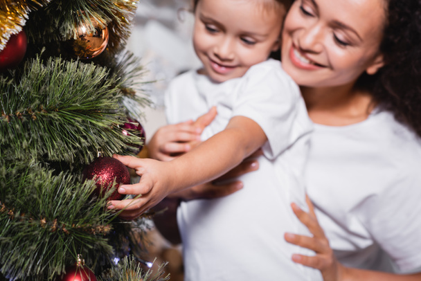 El foco selectivo de la madre y la hija tocando la bola de Navidad en el pino festivo - Foto, imagen