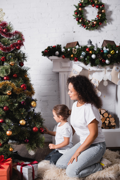 Vista lateral de la madre y la hija sentadas cerca de pino festivo y chimenea en casa - Foto, imagen