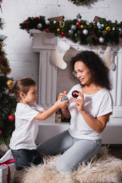 Hija y madre con bolas de Navidad cerca de pino decorado y chimenea  - Foto, Imagen
