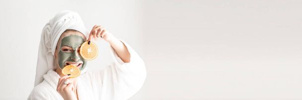 kosmetologia i spa. Młoda kobieta z glinianą maską błotną na twarzy, trzymająca plasterki grejpfruta na białym tle. Długi format banera. - Zdjęcie, obraz